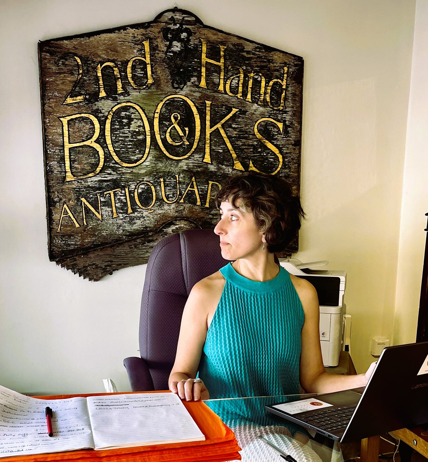 Lucy Ashton Geering in profile sitting at the desk in Voewood Rare Bookshop Holt wearing a halterneck dress by Diesel, with Voewood Rare Books former shop sign on the wall behind her. Photograph by Louisa Young, 01.08.2026. 